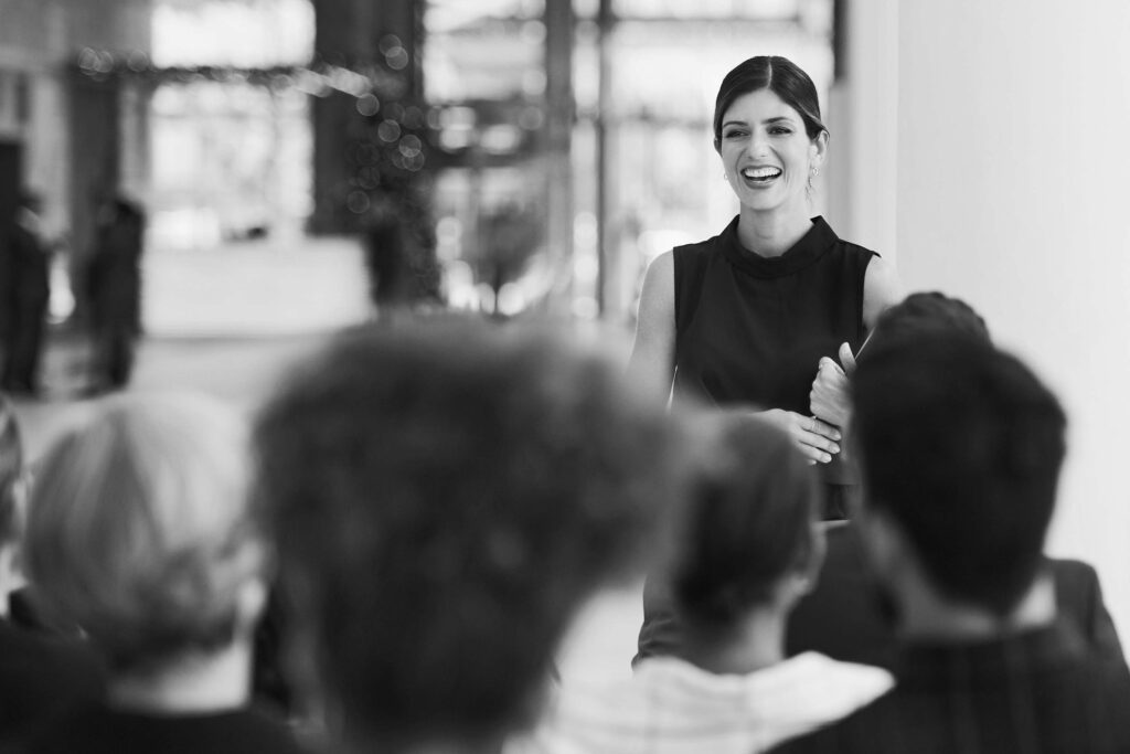 Black and white photo of lady smiling and presenting to a room of people